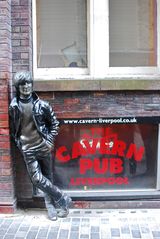 A statue depicting a young Lennon outside a brick building. Next to the statue are three windows, with two side-by-side above the lower, which bears signage advertising the Cavern pub.