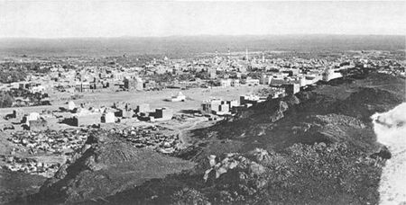 Black-and-white photograph of a city in the desert showing a basaltic ridge on the right and a skyline with numerous buildings among which is a domed mosque with two minarets