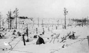 A snowy hill with stone barriers and barbed wire in front, and a distant bunker in behind.
