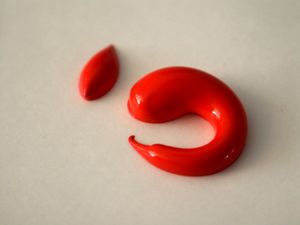 A blob of red acrylic paint shaped like a slug glistens as it rests against a white surface with a small blob to its upper left.