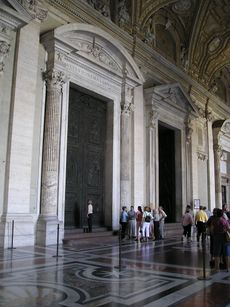 Photo shows view of vestibule with three huge doorways leading to the church's interior. The doors are framed by columns and have pediments. The floor is of inlaid marble. The nearest doorway is closed by two huge ancient bronze doors. A group listens to a tour guide while one woman examines the doors.
