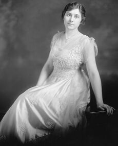 A slightly built, dark haired woman wearing a white dress and leaning backward onto her left arm