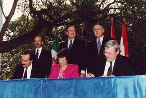 Two men and one women sit at a table and sign a piece of paper, while three men in suits stand behind them, in front of a set of limp flags