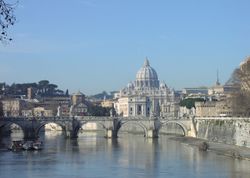 A view of Rome on a sunny afternoon looking along the river. A bridge crosses the river and beyond it is a hill on which the grey dome of St. Peter's rises above ancient buildings and dark pine trees.