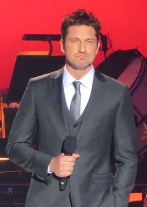 Gerard Butler standing with a microphone with a red background in a suit and tie facing the camera