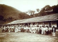a large group of men, women and children holding agricultural implements and standing in front of a long, low building with hills rising in the background