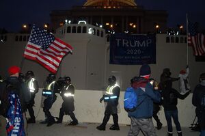 A photo taken near dusk of the Capitol building. A large Trump flag is hung on a railing. Protestors are still remain, and a small line of police officers yield shields.