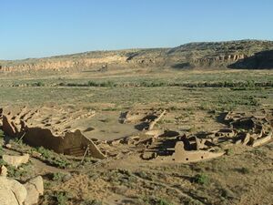 Daytime view looking down on a desert valley: in the near distance, a large semi-circular set of tumbled-down and ruined walls, greyish-yellowish brown in color. The far side of the ruins is a straight line, running left-right, roughly parallel to a line of cliffs in the far distance.