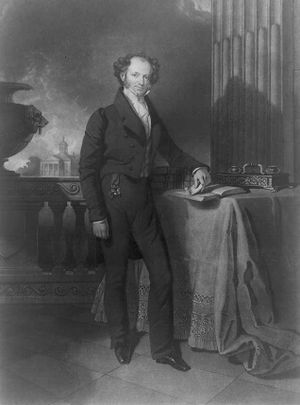 Engraved full-length portrait of a balding man standing next to a table with his left arm resting on a book and in the background a stone balustrade beyond which are trees and a building with columned portico