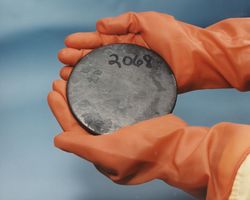 Two hands in brown gloves holding a gray disk with a number 2068 hand-written on it