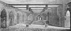 A monochrome illustration of a stone and brick-walled room. An open doorway is to the right. The left wall contains equally spaced arches. The right wall is dominated by a large brick arch. Three arches form the third wall, in the distance. The floor and ceiling is interrupted by regularly spaced hexagonal wooden posts. The ceiling is spaced by wooden beams.