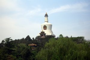 Color photograph of a white, bell-shaped building composed (من أسفل إلى أعلى) of a square base, three round disks of increasingly smaller diameter, a cut reverse cone, and a thinner tapering column with horizontal flutings crowned by the golden statue of a sitting figure. It appears to emerge from a forested area, against the background of a slightly cloudy blue sky.