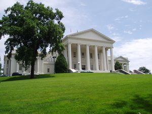 All white Neoclassical building with pediment and six columns rises on a grassy hill with a large American elm tree in the left foreground. Two boxier, but similarly styled wings are attached at the building's rear.