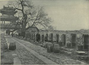 A black-and-white picture of a stone-paved alley going from bottom right to top left leading to a three-roofed gate and bordered on the right by a line up of small roofed cubicles open on one side.