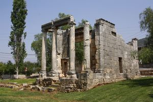 Three quarters view of a temple with four standing ionic columns at the front; the temple is built with gray limestone and stands on a green meadow with a poplar tree to its left.