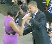 The Obamas face each other and bump fists on stage. She wears a purple dress and he wears a dark suit. Several signs read "CHANGE WE CAN BELIEVE IN" and several photographers take photos.