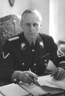 Portrait of a middle-aged man with short grey hair and a stern expression. He wears a dark military uniform, with a swastika on one arm. He is seated with his hands on a table with several papers on it, holding a pen.