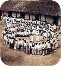 Photograph showing a group of people dressed in white, who have gathered in front of a tile-roofed farm building and observe another large group which has formed a large circle surrounding 5 men straddling large drums, a woman and 2 other men.