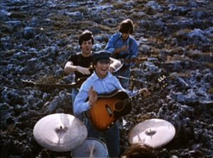 The Beatles performing music in a field. In the foreground, the drums are played by Starr (only the top of is head is visible). Beyond him, the other three stand in a column with their guitars. In the rear, Harrison, head down, strikes a chord. In the front, Lennon smiles and gives a little wave toward camera, holding his pick. Between them, McCartney is jocularly about to choke Lennon.